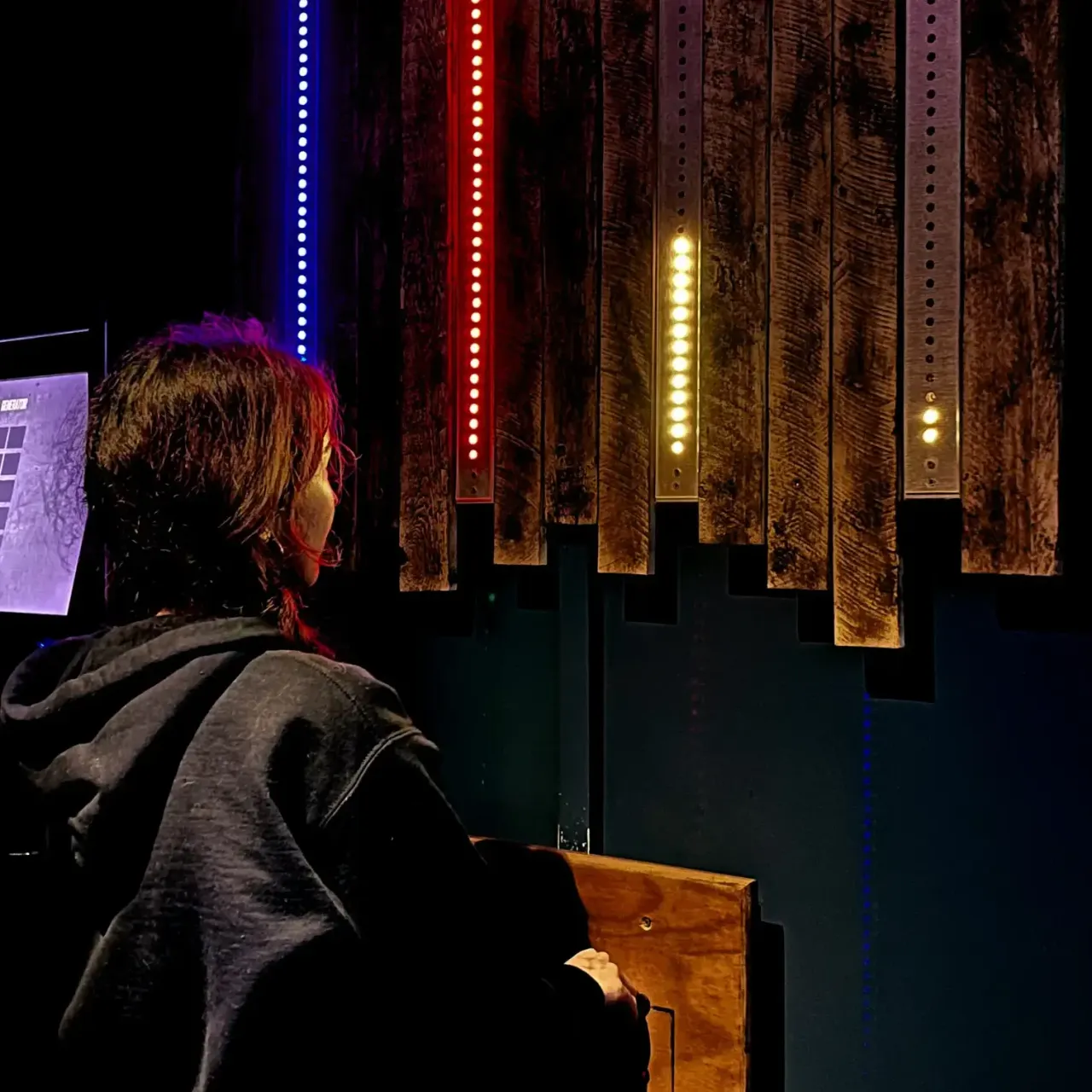 Person viewed from behind sitting at a desk in a darkened escape room with illuminated brick walls and coloured LED light strips.