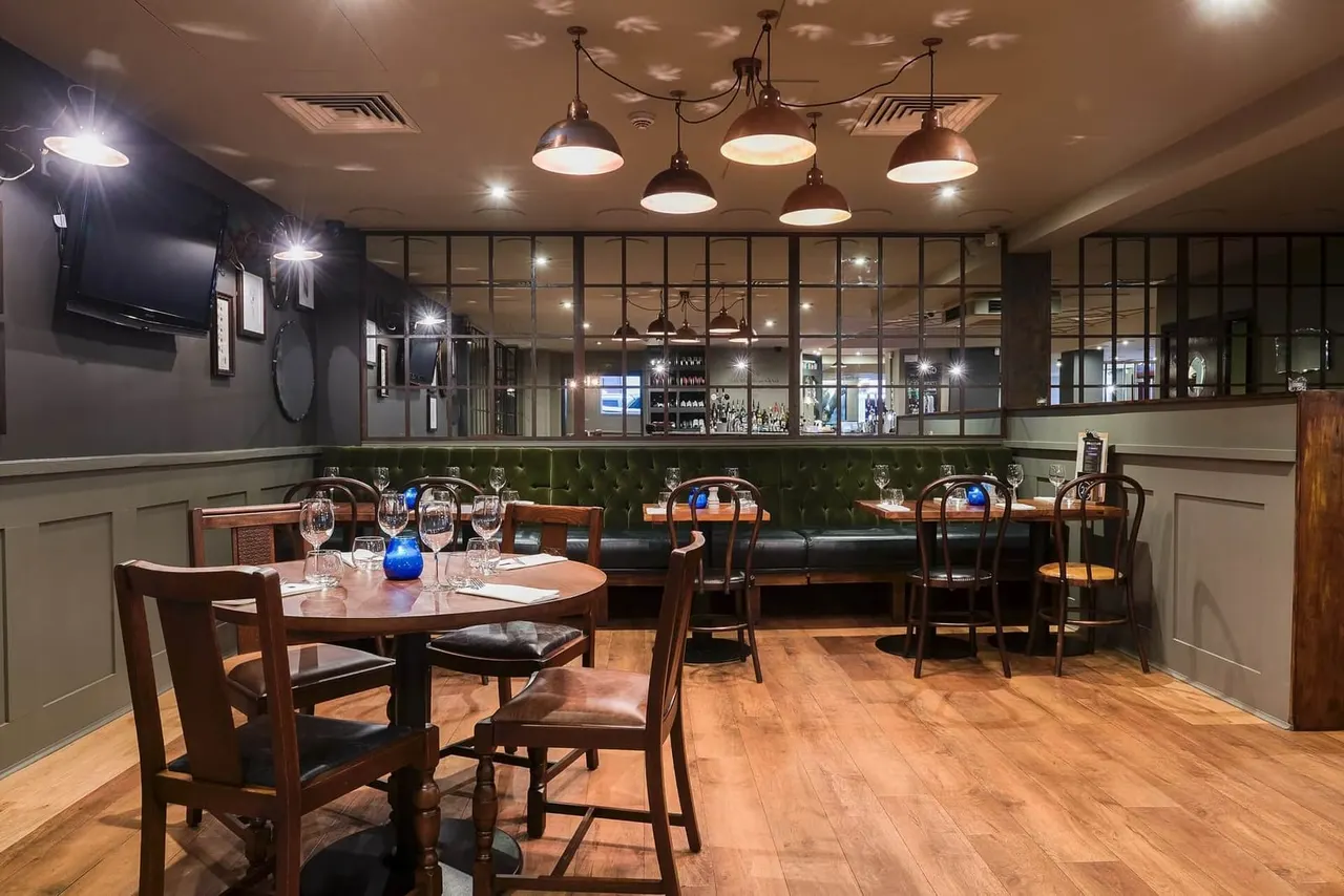 Empty restaurant dining room with wooden tables and chairs, green leather banquette seating, and industrial pendant lighting.