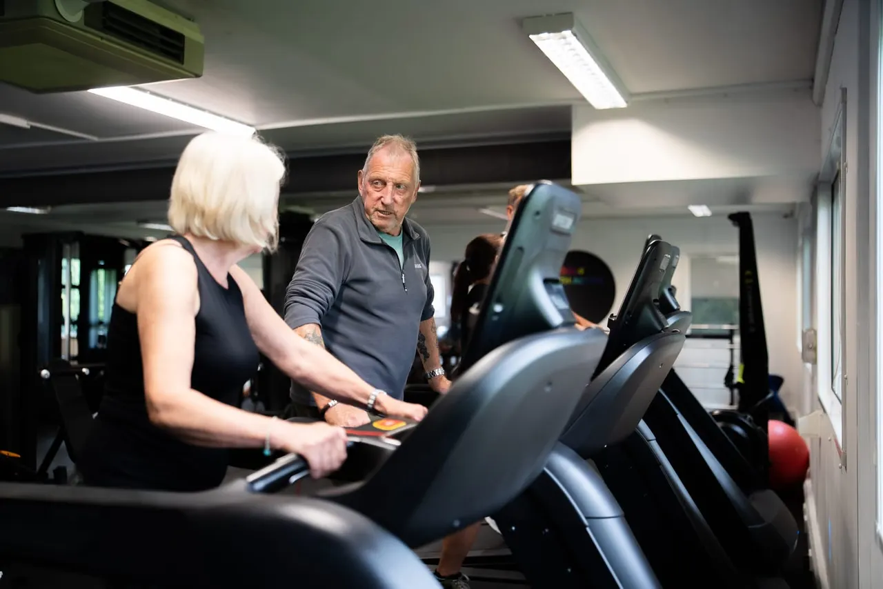Older woman and man using exercise bikes in a modern gym facility with other visitors in the background.
