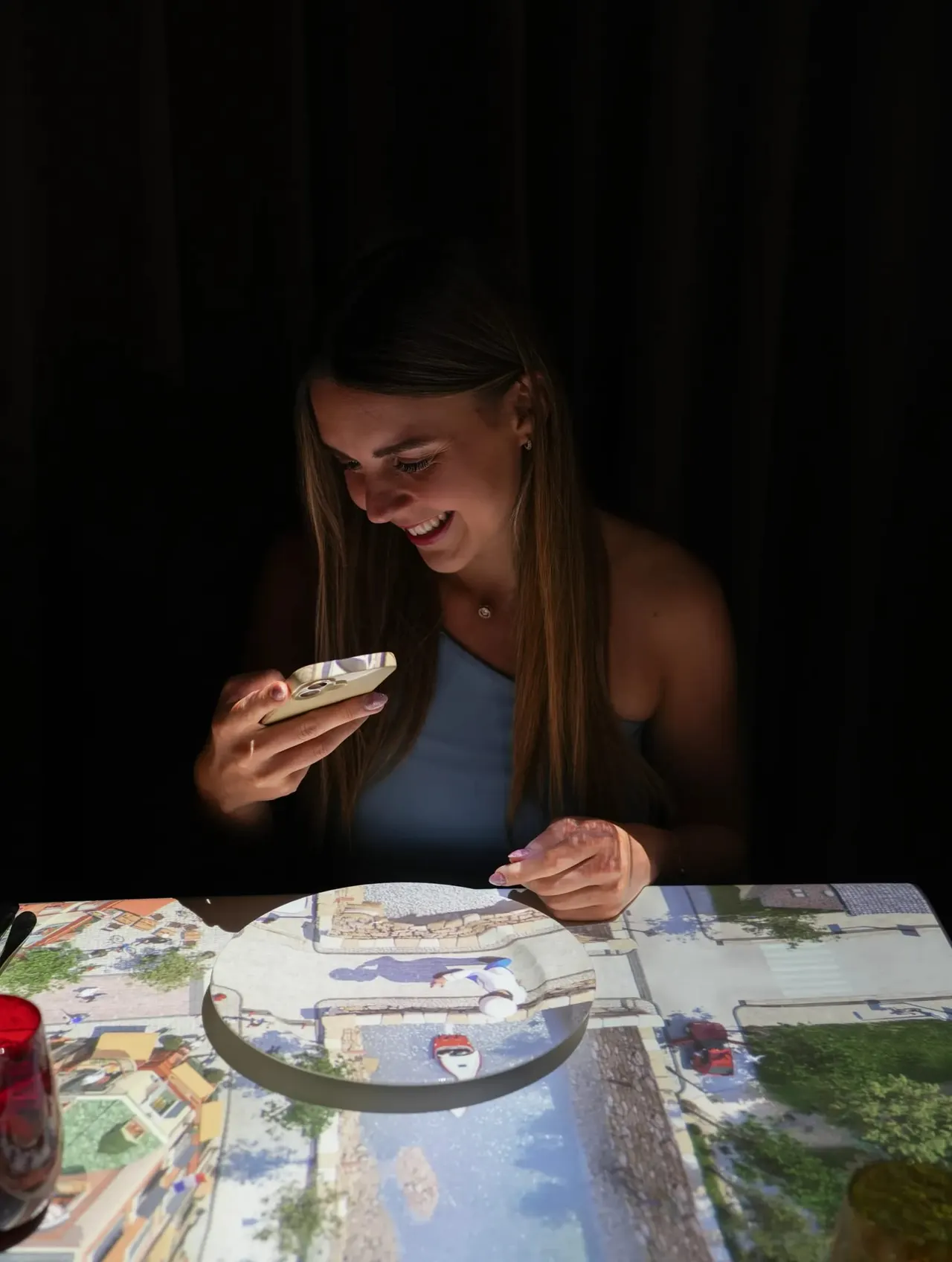 Woman smiling whilst using a smartphone to view an interactive miniature dining scene on a plate.