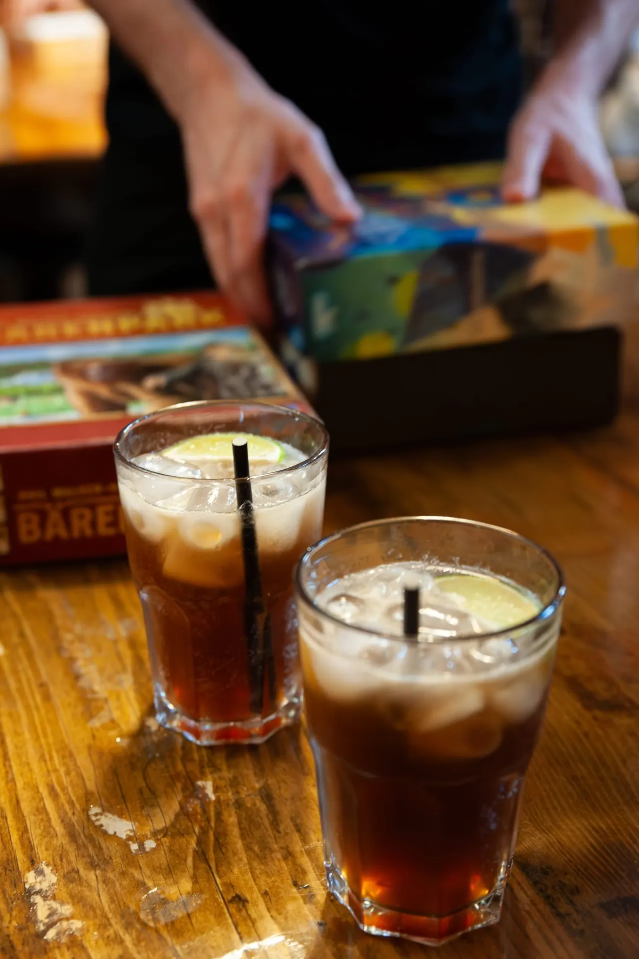 Two glasses of cola with ice and lime on a wooden table, with people's hands playing board games in the background.