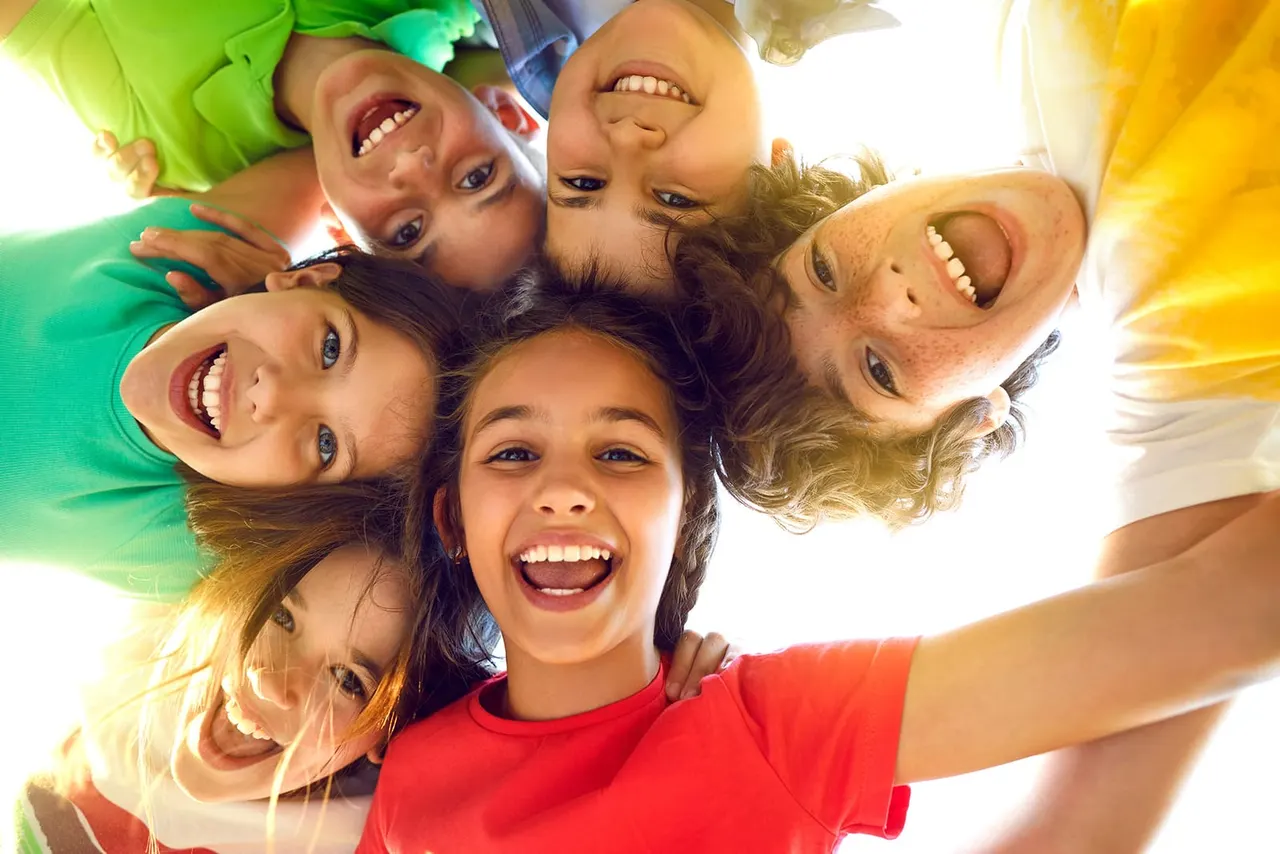 Group of smiling children and adults huddled together looking down at camera in bright sunshine.
