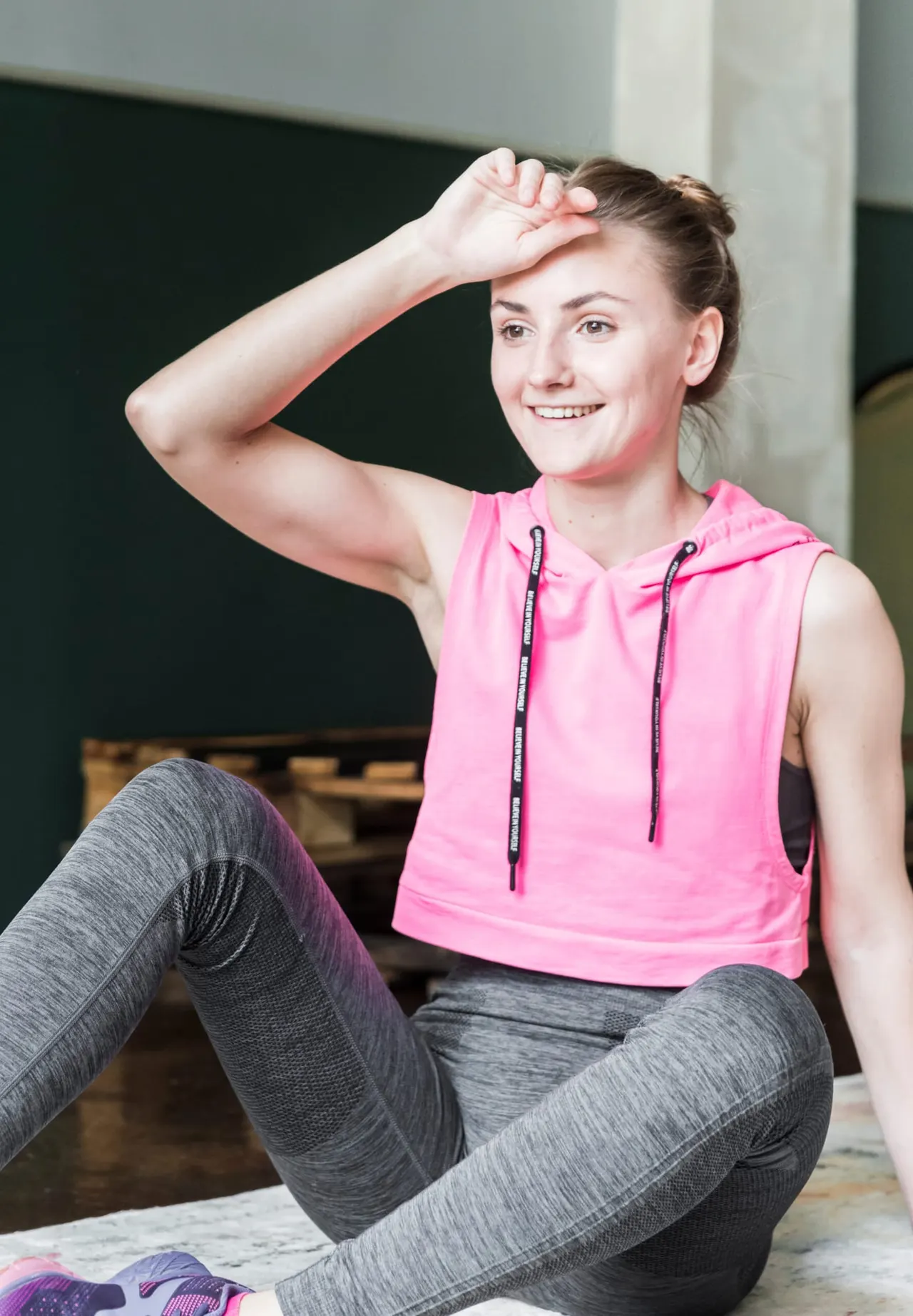Woman in pink sports top and grey leggings smiling while posing in a fitness studio.
