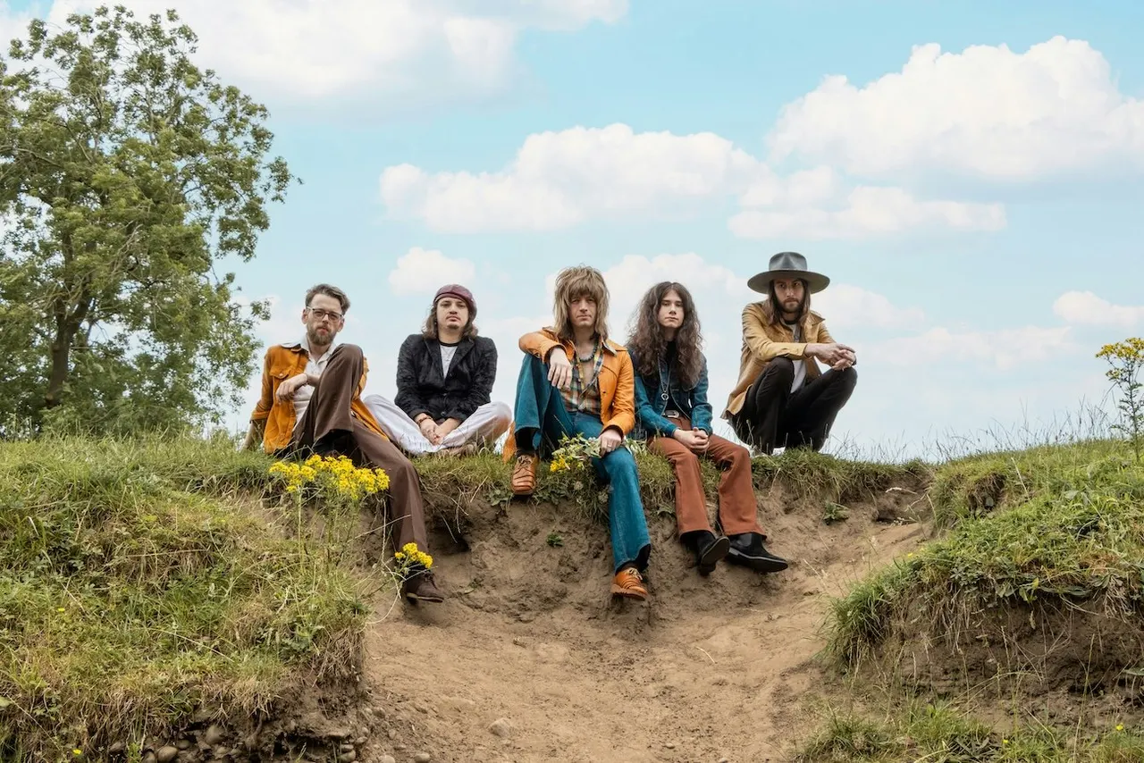 Five people sitting on a dirt bank in countryside, wearing colourful vintage-style clothing against a blue sky with clouds.