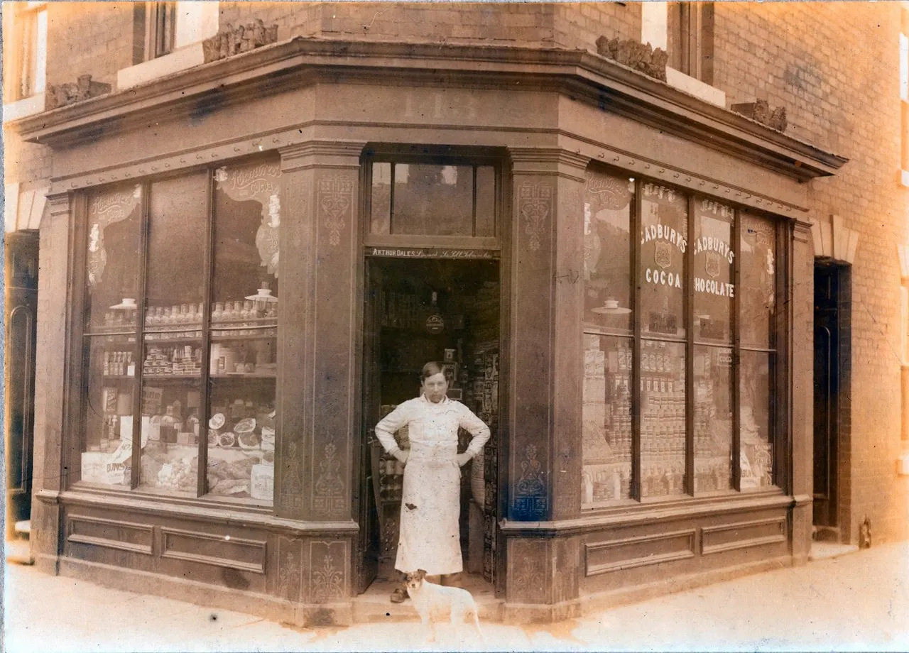 Woman standing in doorway of vintage corner shop with display windows advertising Cadbury's cocoa and chocolate.