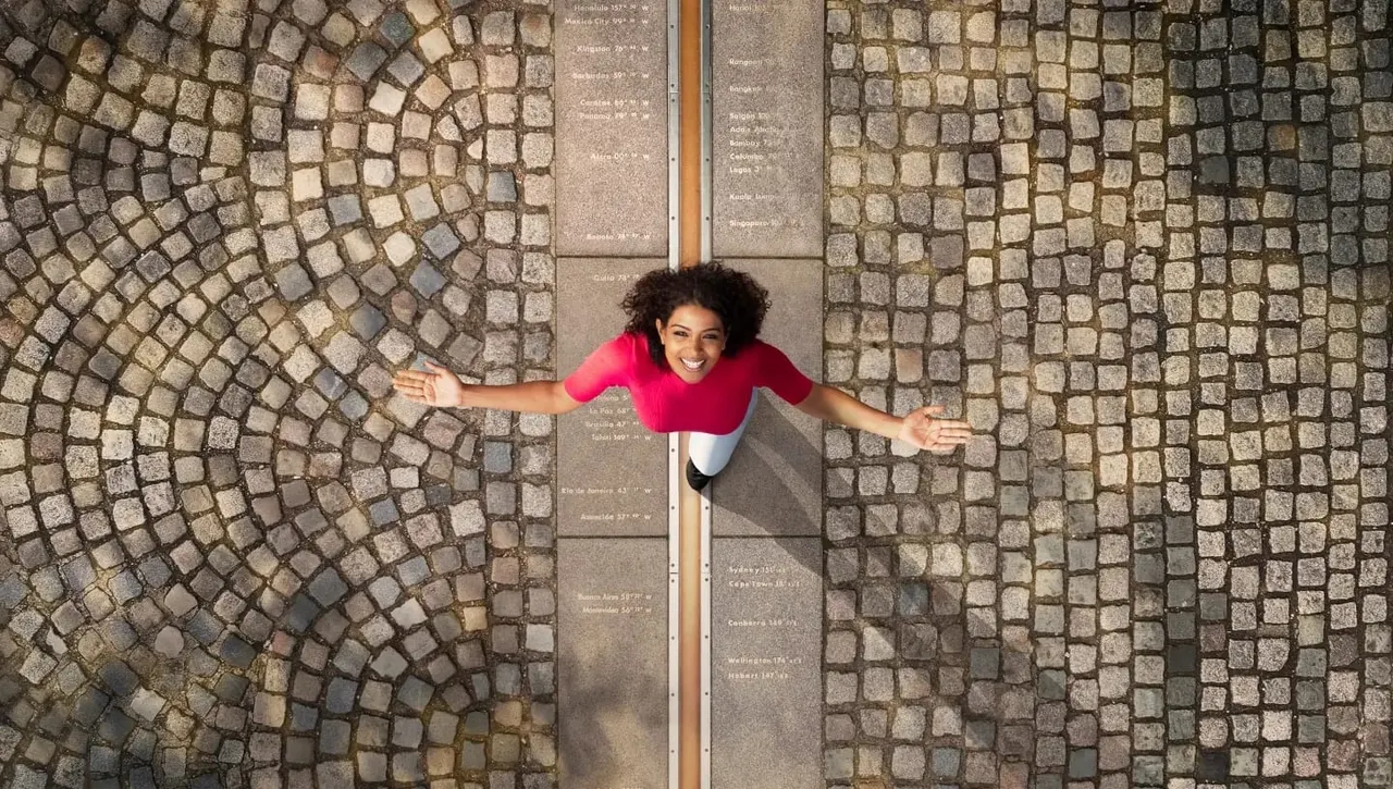 Woman in bright pink top lying on cobbled ground at Greenwich meridian line, arms outstretched, smiling at camera.