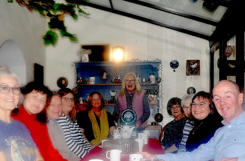 A group of people smiling at the camera inside a decorated coffee shop with blue display cabinet and hanging plants.