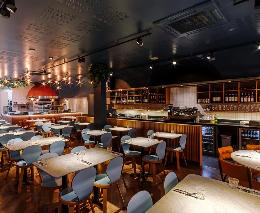 Modern restaurant interior with dining tables, bar counter with wine bottles, and open kitchen with red pizza oven.