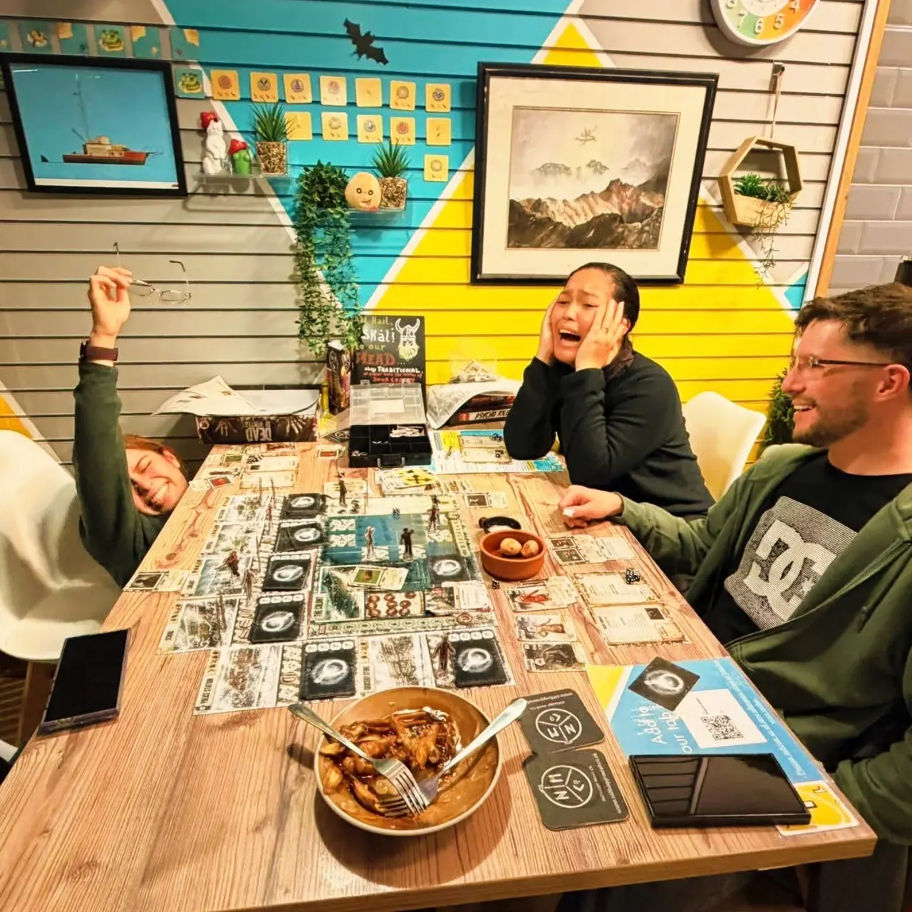 Three people playing a board game at a wooden table with food and drinks in a colourful venue.