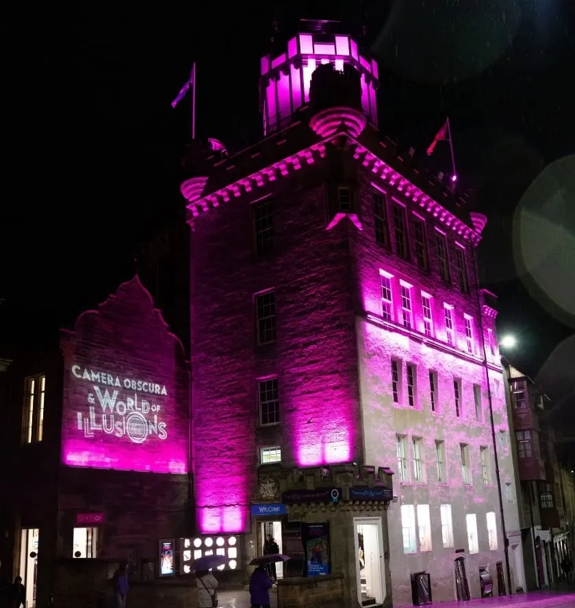 Historic stone tower building illuminated in bright magenta at night, with signage for Camera Obscura and World of Illusions visible.