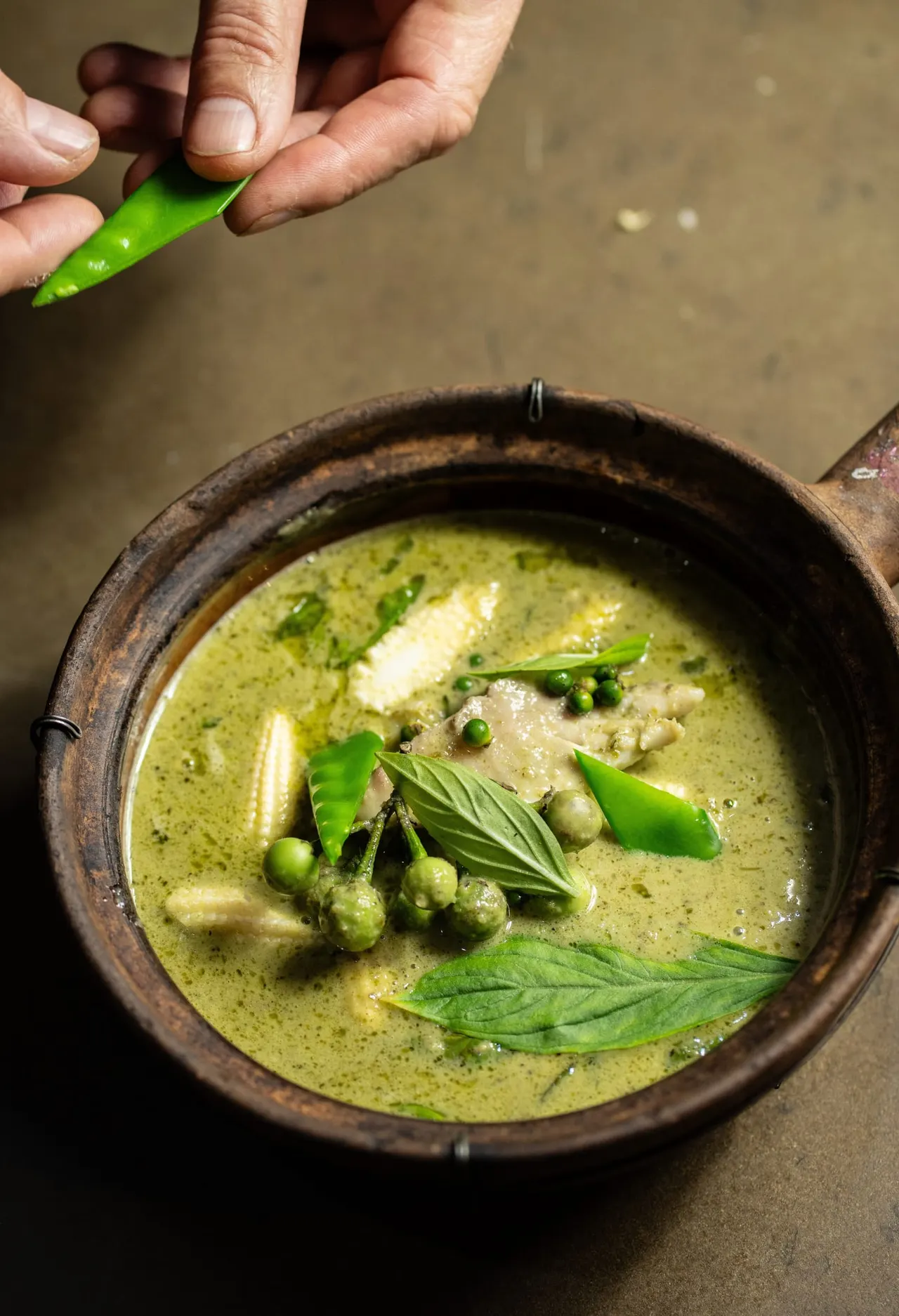 Person holding a green chilli above a rustic bowl of green curry with peas, herbs and coconut broth.