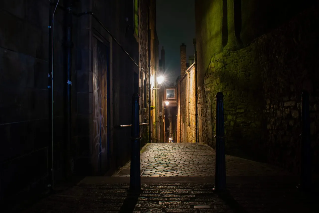 Narrow cobbled alley at night with historic stone buildings lit by street lamps and a doorway ahead.