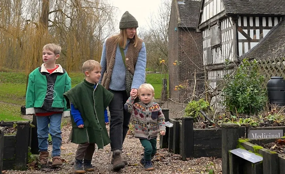 Woman and three young children walk through a historic garden with raised beds beside a half-timbered Tudor building.