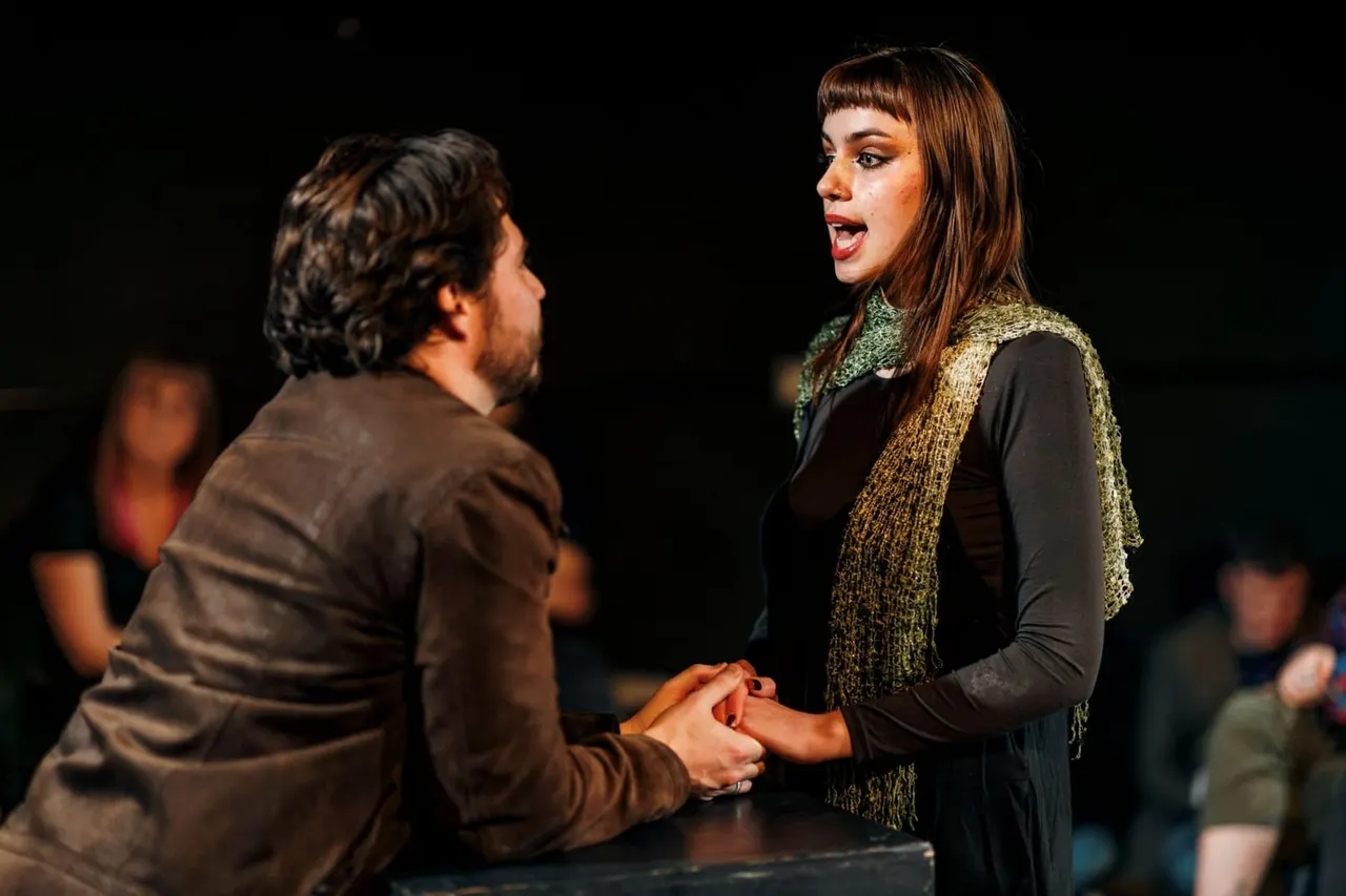 Two actors performing a scene on stage, holding hands whilst facing each other against a dark background with audience members visible.