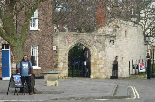 Person standing with information boards outside a historic stone gatehouse with arched entrance and bare winter trees.
