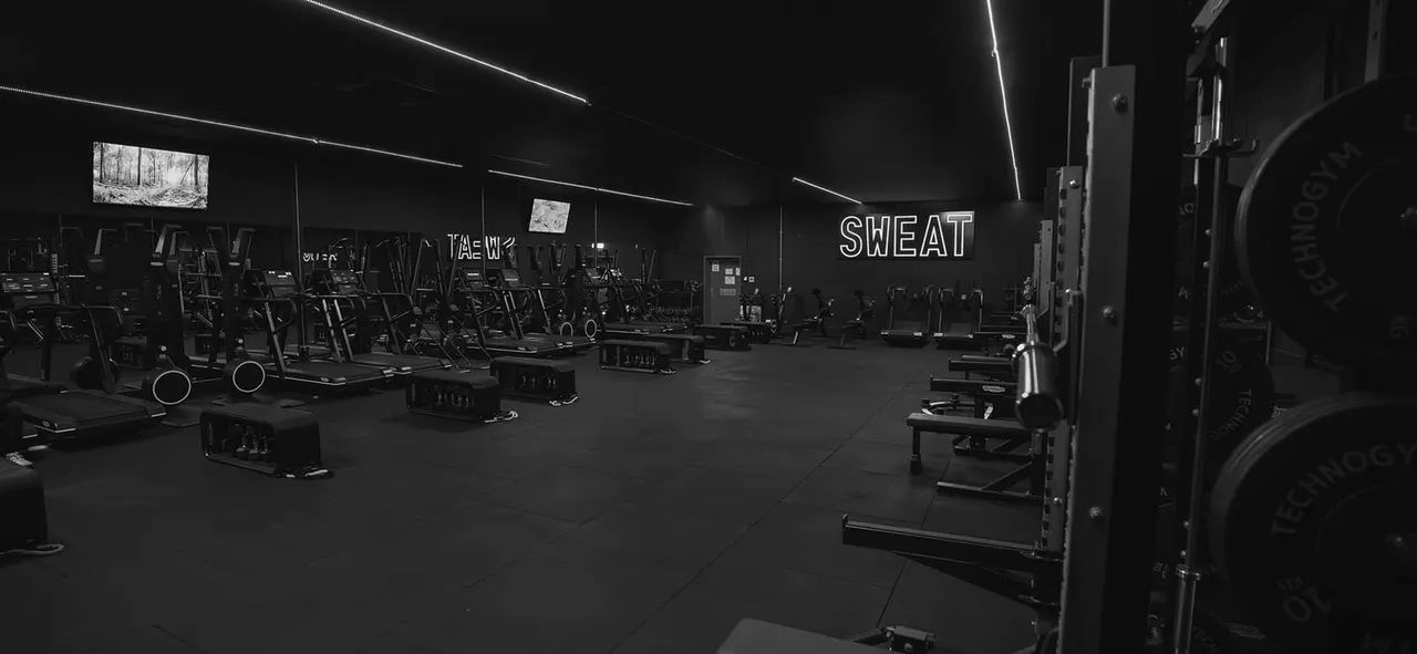 A spacious gym interior with rows of exercise equipment including treadmills and weight machines, illuminated by overhead lighting with neon "SWEAT" signage.