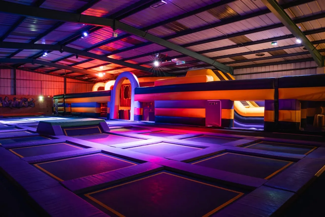 Brightly lit indoor trampoline park with neon-illuminated bounce platforms and orange inflatable structures under a colourful ceiling.
