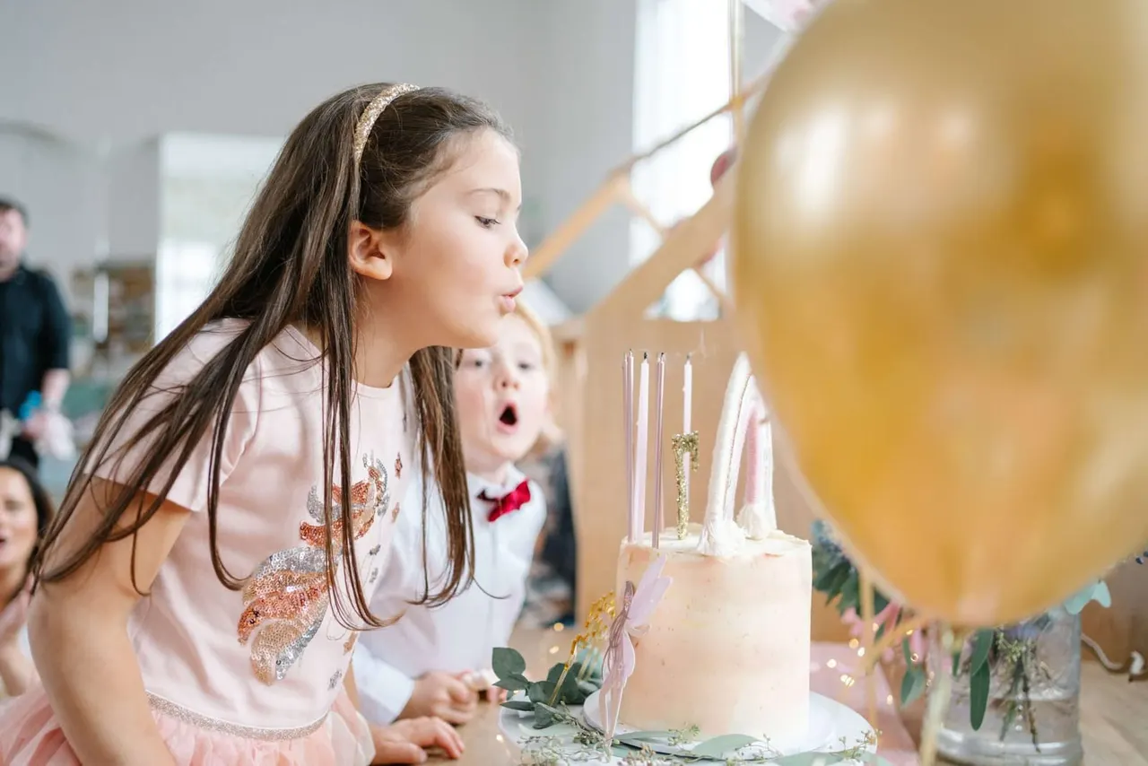 Child blowing out candles on a birthday cake at an indoor party celebration with other children watching.