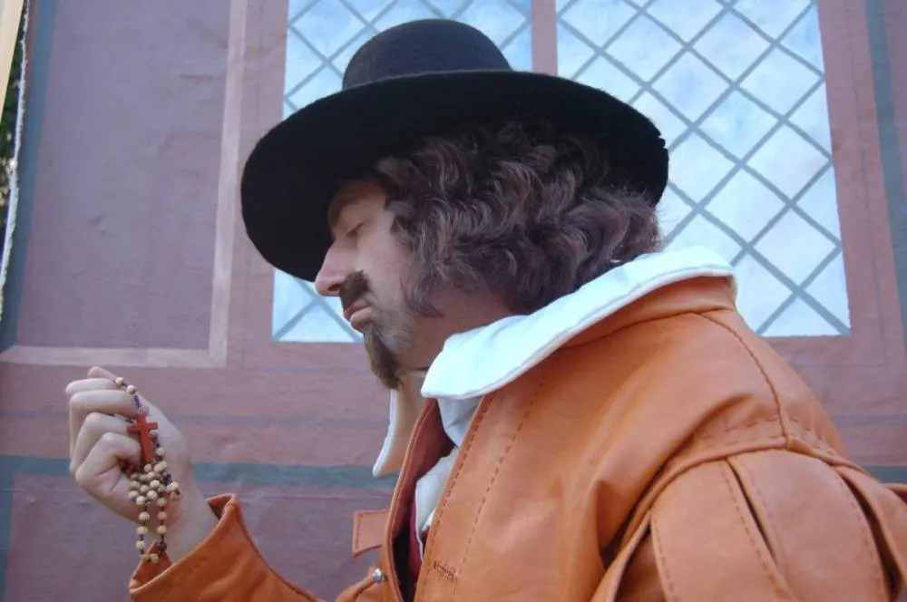 A wax figure of a bearded man in period costume holding a rosary, wearing a black hat and orange-brown jacket.
