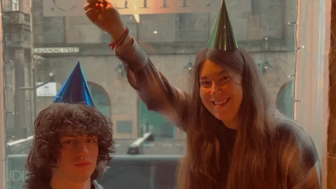 A young woman wearing a green party hat raises her fist in celebration whilst posing with a statue wearing a blue party hat.