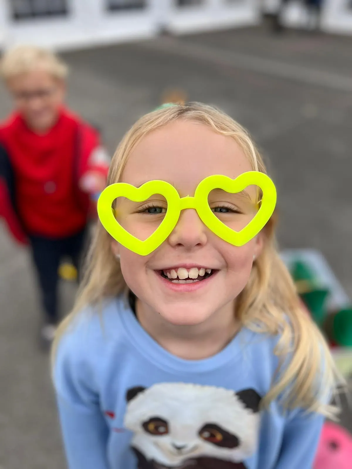 Young child wearing neon yellow heart-shaped glasses smiles at camera in indoor play area.