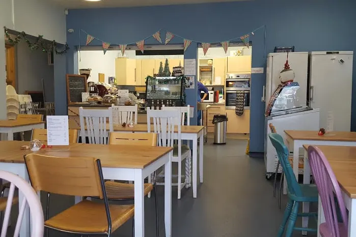 A café interior with dining tables and chairs, a serving counter with kitchen appliances, and festive decorations on blue walls.
