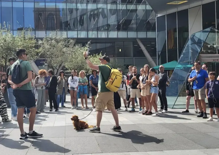 A man with a small brown dog addresses a large crowd gathered in a sunny city square outside a modern glass building.