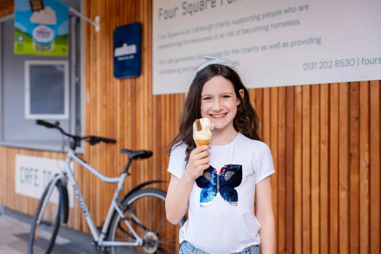 A smiling girl holding an ice cream cone stands in front of a wooden wall inside a café, with a bicycle visible to the left.