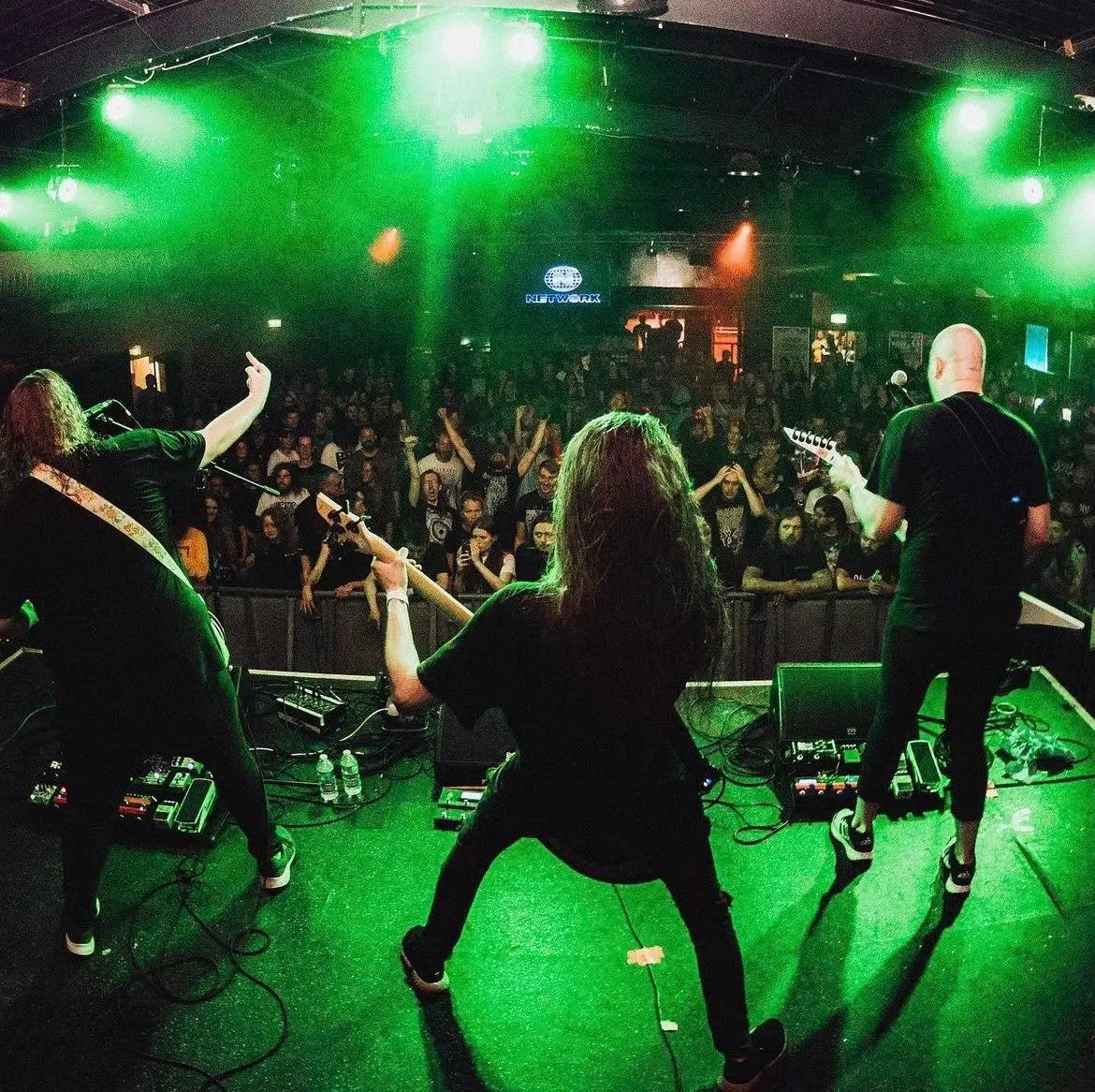 A live band performs on stage under bright green lighting to a crowded audience in an indoor music venue.