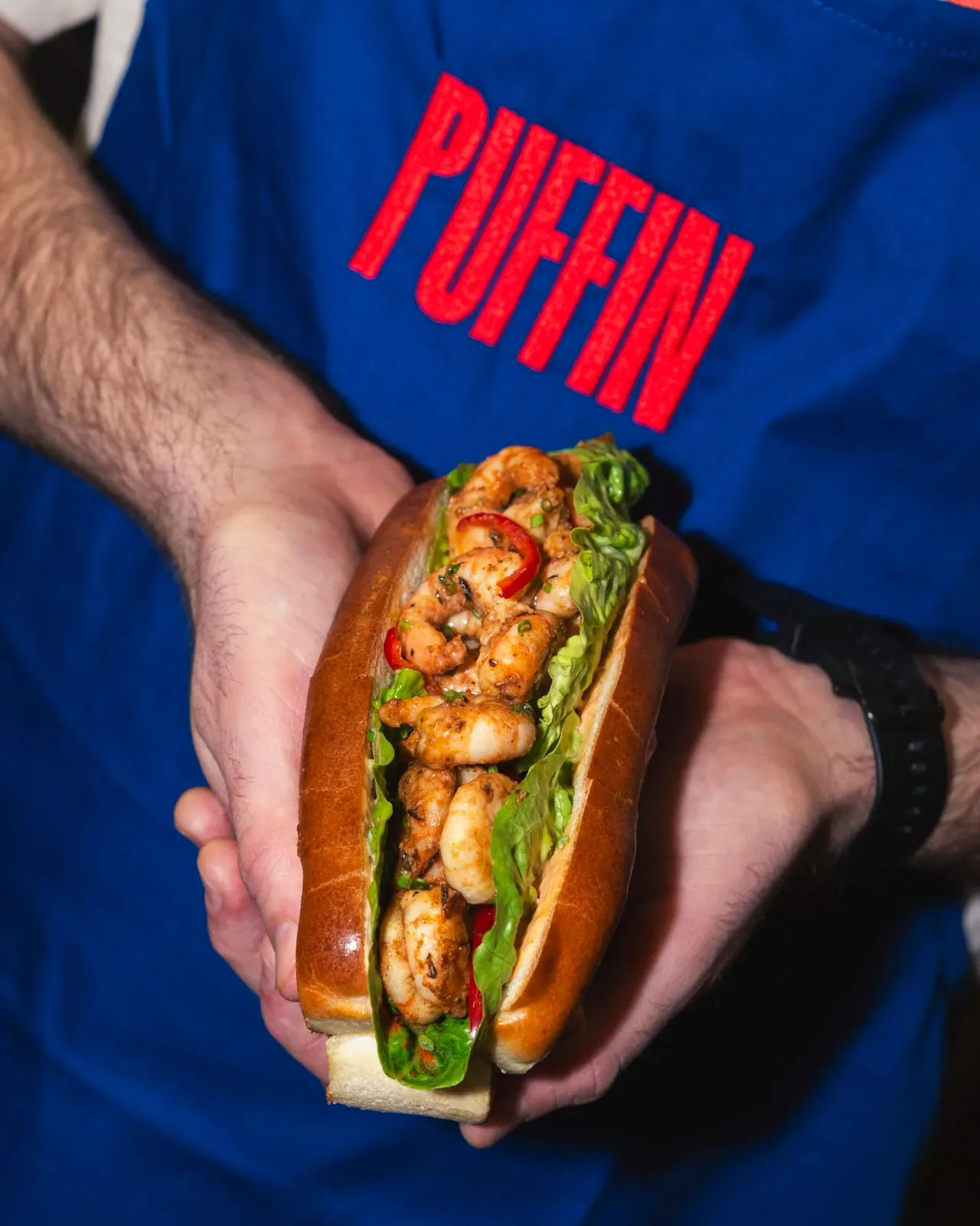Staff member in blue uniform holding a cooked prawn sandwich with lettuce and red pepper.