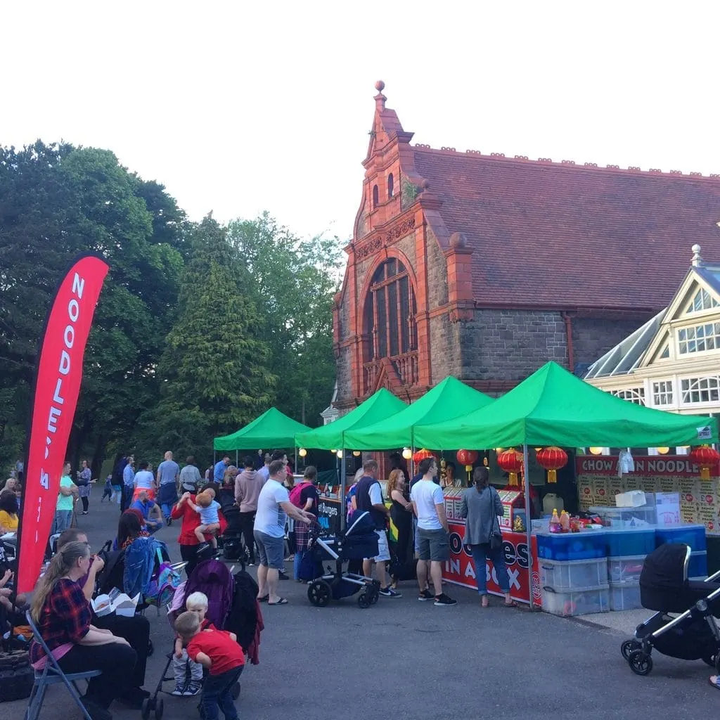 Outdoor festival or food market with green marquees, food stalls, and crowds gathered in front of a red-brick church building.