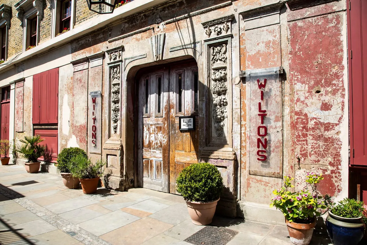 Historic building facade with weathered red and cream painted walls, ornate stone details, dark wooden doors, potted plants, and red signage reading "Wiltons" on a sunny day.