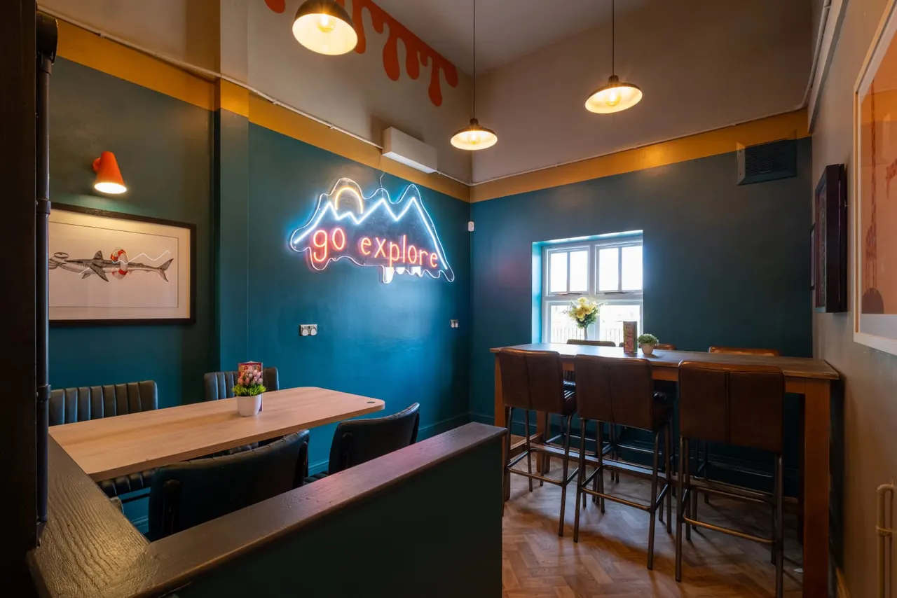 A modern dining space with teal walls, wooden tables and dark chairs, illuminated by pendant lights and a neon "go explore" sign.
