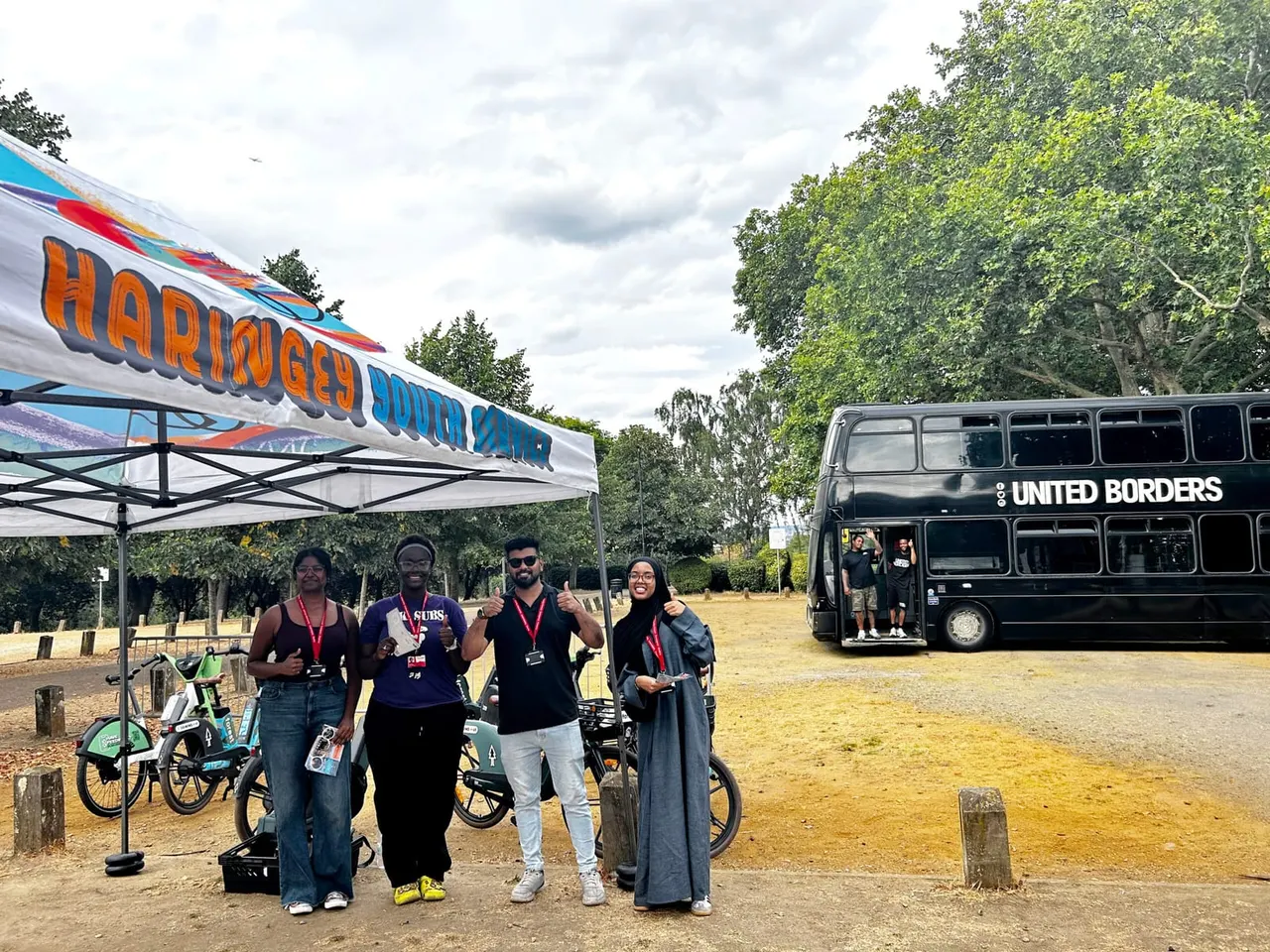 Four people stand together with bicycles beneath a marquee at an outdoor event, with a double-decker bus nearby.