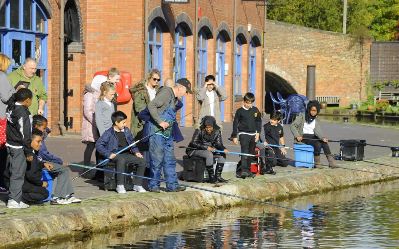 Children and adults fishing together on the towpath by a canal, with a brick building behind them.