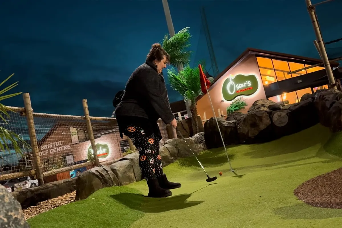 Woman playing mini golf at an indoor course with bright green astroturf and tropical decorations.