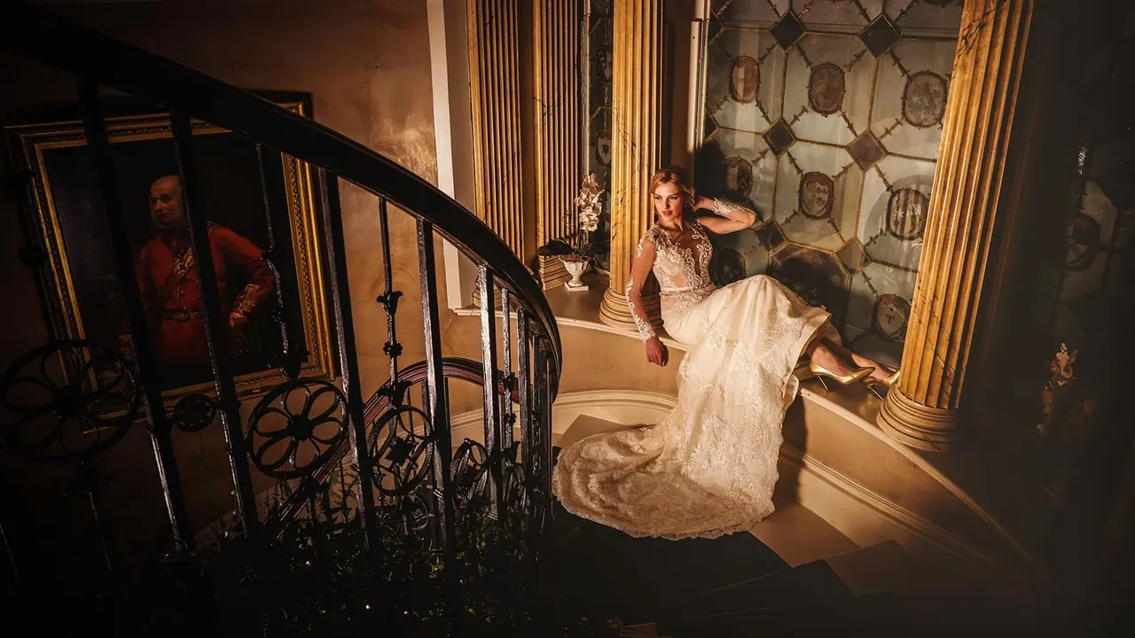 Woman in an ornate ivory wedding dress posing on a grand staircase in a historic mansion interior.