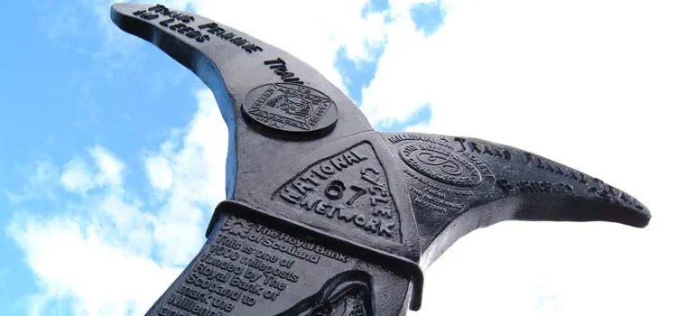 Large black metal whale tail sculpture against blue sky with white clouds.