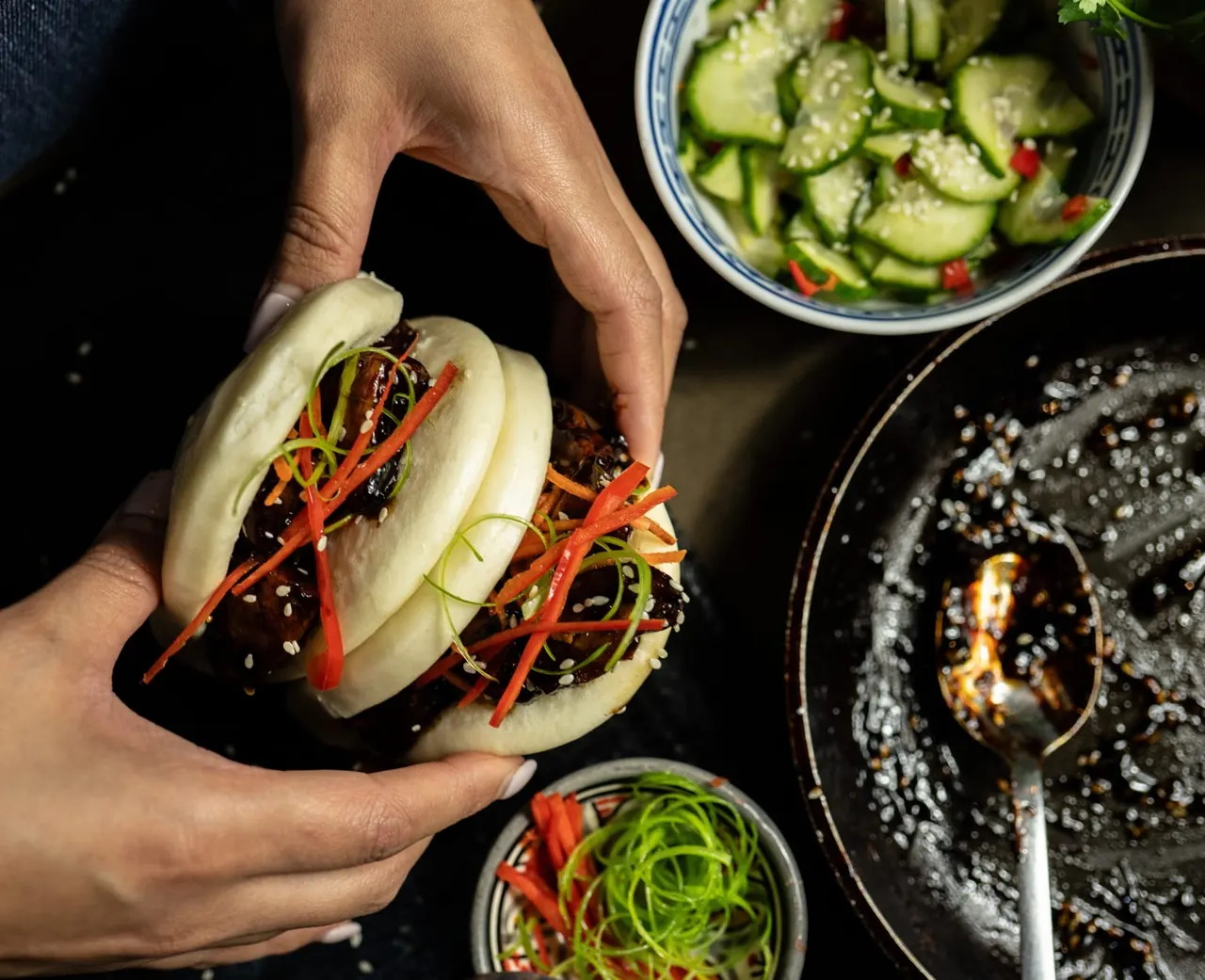 Person holding a steamed bun filled with braised meat and red pepper strips, surrounded by bowls of garnishes and condiments.