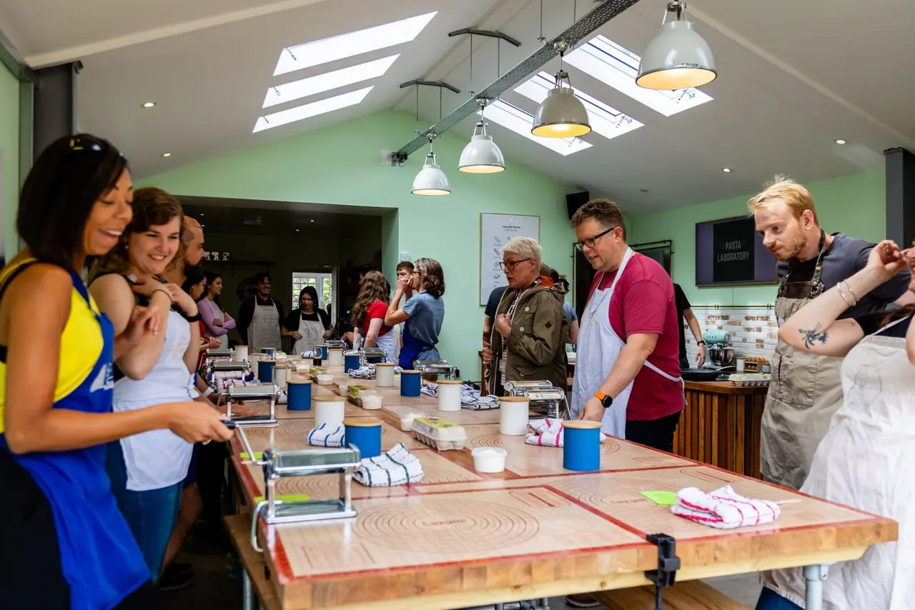 A group of participants in a bright, modern kitchen class with green walls and skylights, standing around wooden work tables during a pasta-making activity.