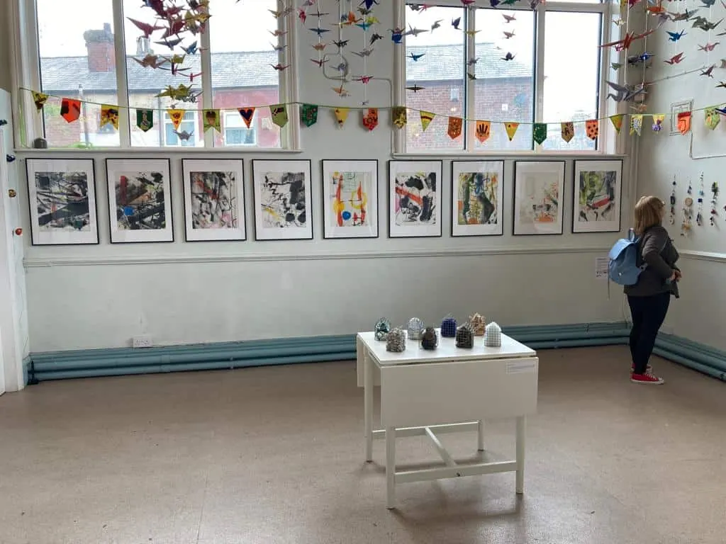 Child viewing artwork in a bright gallery space with framed prints on walls, hanging paper birds and bunting decorations, and sculptural objects on a white plinth.