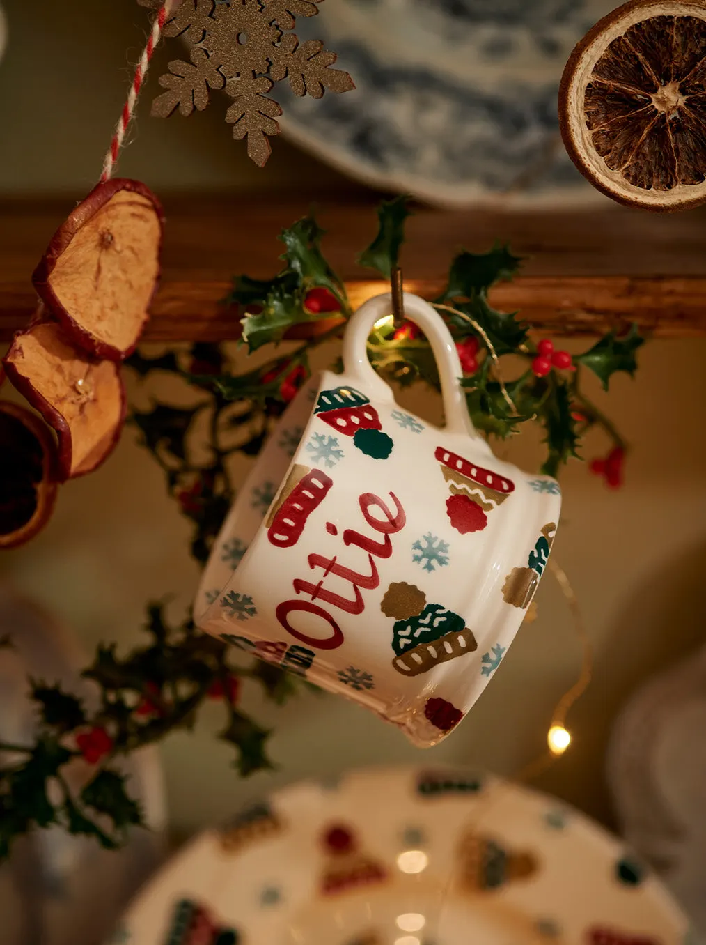 Festive ceramic mug ornament hanging among dried citrus slices, holly sprigs and snowflake decorations on wooden surface.