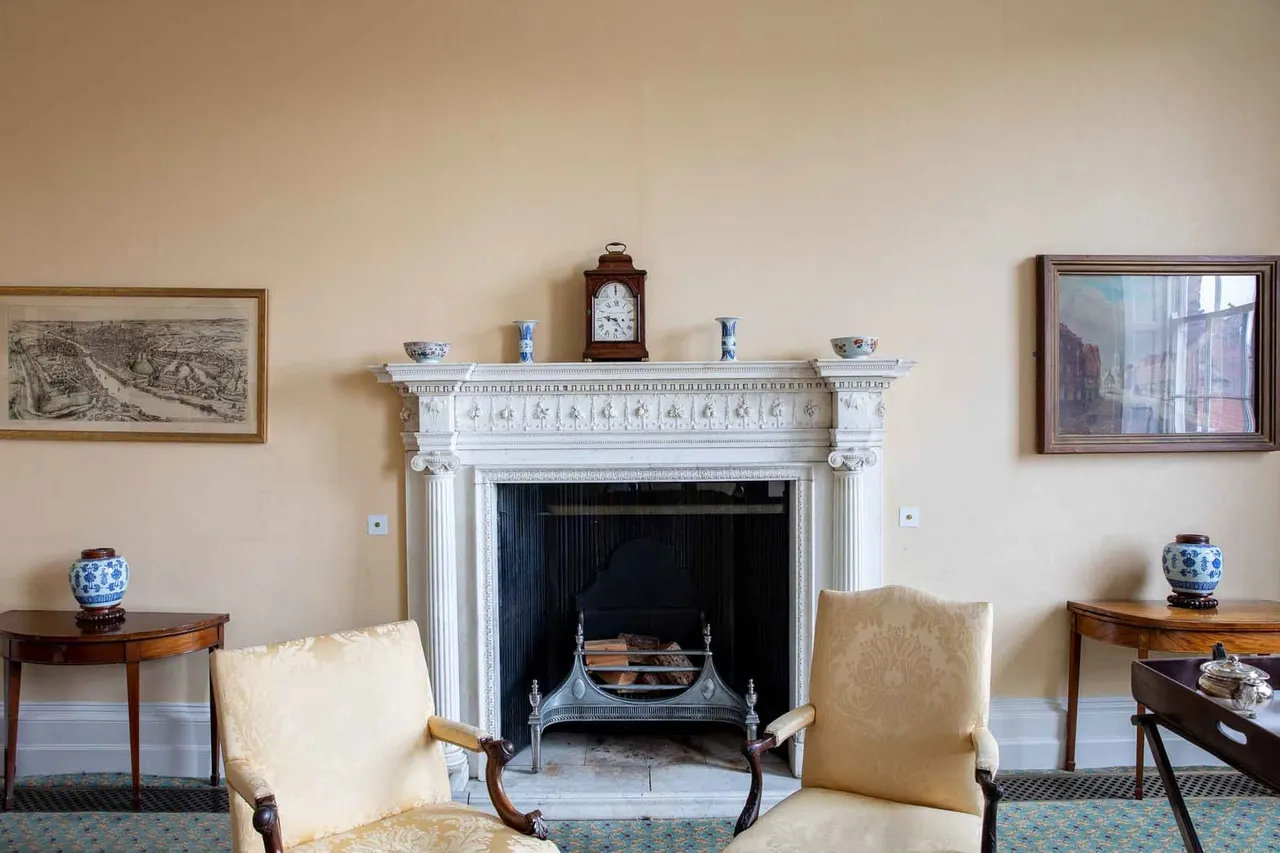 A formal Georgian sitting room with a white marble fireplace, two cream upholstered chairs, wooden side tables with blue and white porcelain, framed artwork on cream walls, and period furnishings.