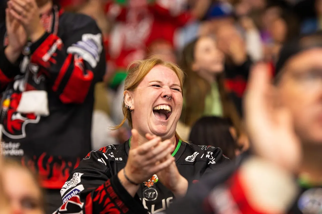 Woman in black and red sports kit laughing and clapping among a crowd of spectators at an indoor venue.