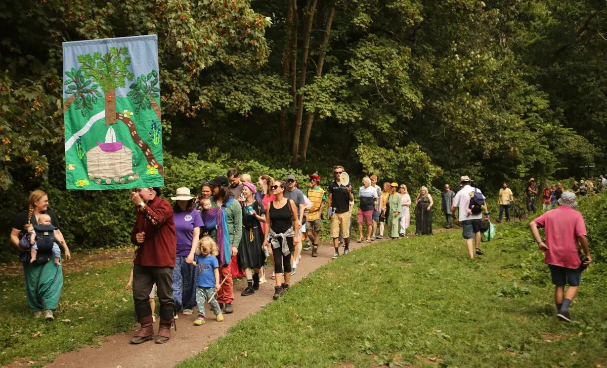 A large group of visitors of mixed ages walking along a woodland path beside a green banner illustration, surrounded by trees and grass.