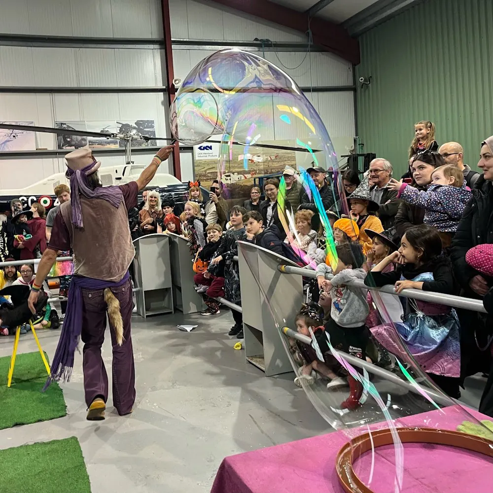 A performer in purple creates a giant iridescent bubble while an audience of children and adults watch in an indoor hangar.