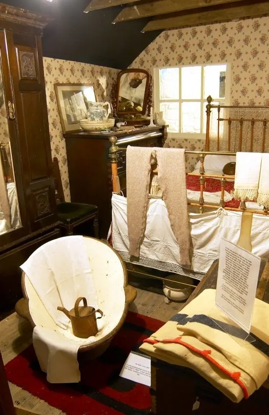 Historic bedroom display with period furniture, floral wallpaper, bathing equipment and domestic artefacts arranged as museum exhibition.