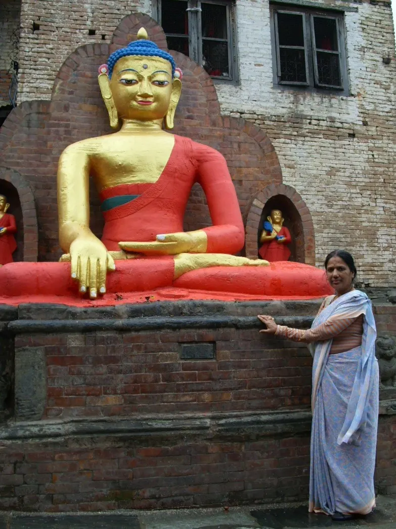 Woman posing beside a large gilded Buddha statue wearing red robes against a brick temple building.