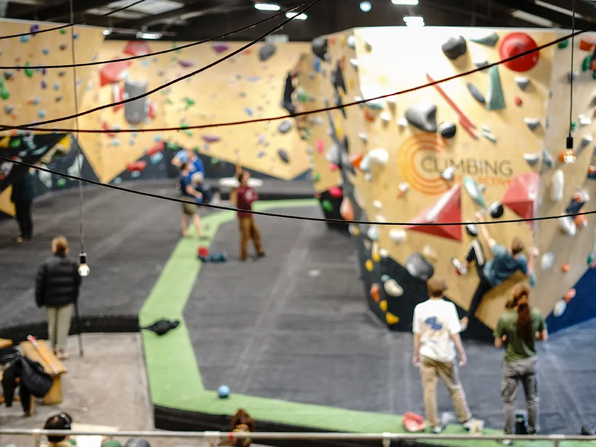 Indoor climbing wall facility with multiple participants climbing coloured holds on tan-coloured walls, with ropes and safety equipment visible.