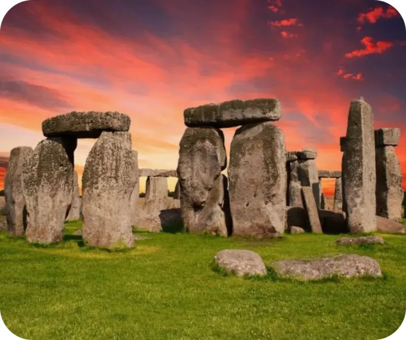 Ancient stone monument with standing megaliths and lintels on grass beneath a dramatic red and orange sunset sky.